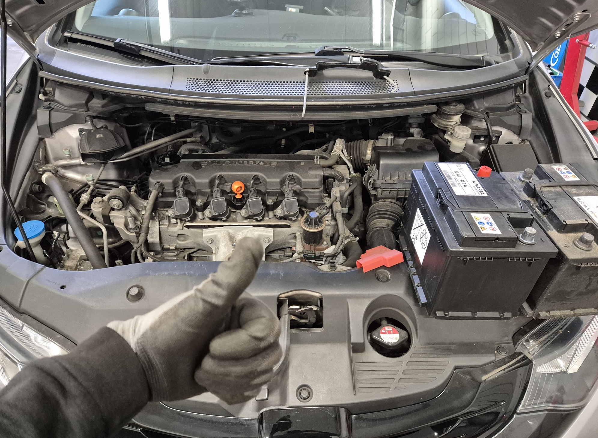 Motorruimte van een Honda met de accu, en een duim omhoog in een werkhandschoen